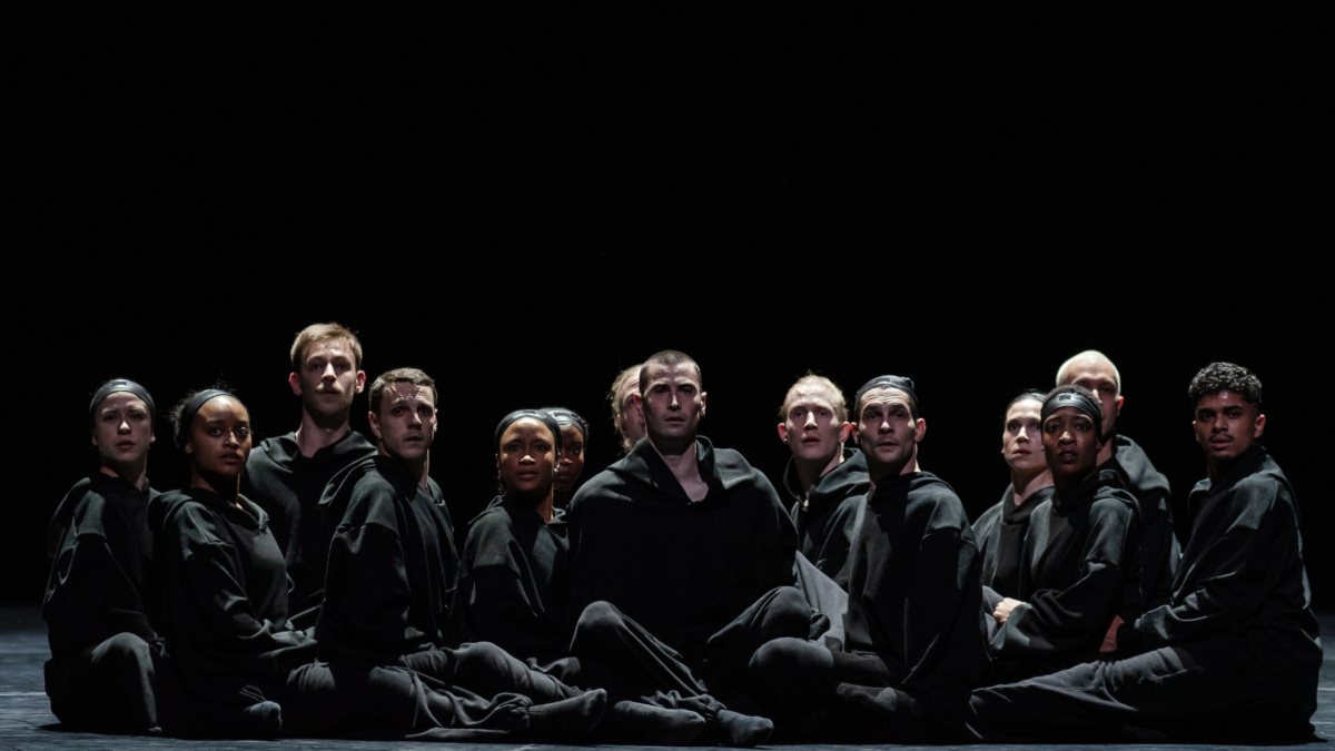 Group of people sitting cross-legged on stage wearing all black looking out.