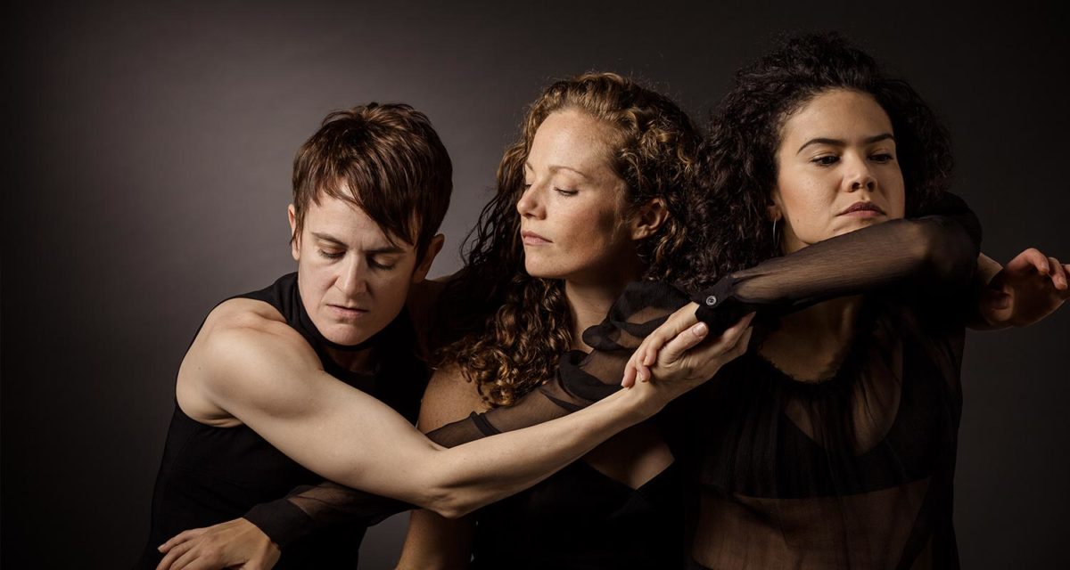 Gibney Company Artistic Associates Amy Miller, Katie Lake, and Zui Gomez. The photo is from the waist up and the dancers' arms intertwine as they lean on one another. 