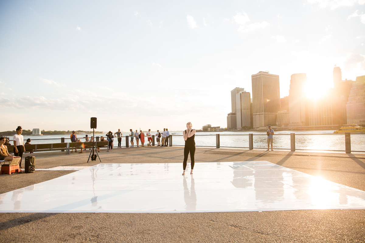 A white dancer standing on a marley floor in front of a New York City skyscape and river with the sun beaming behind them.