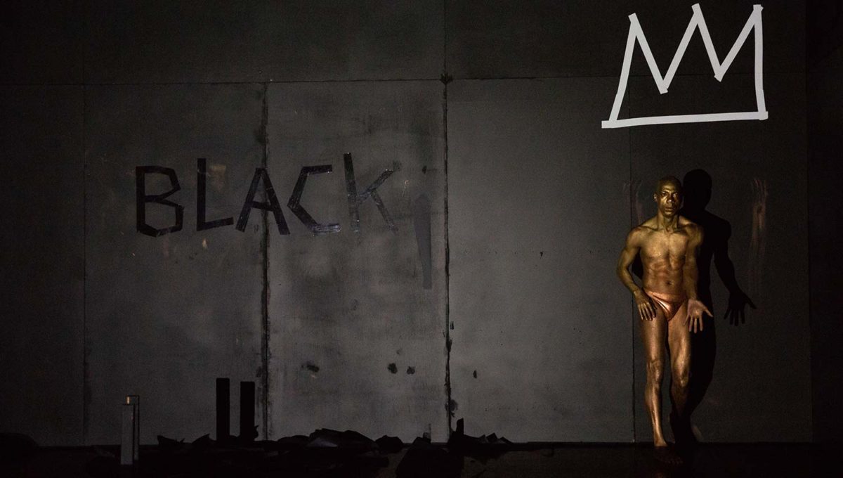 Gibney Company Curation. Photo of Shamel Pitts standing in front of a black wall with the word "BLACK" spelled out in black tape and an image of a crown in white tape. 