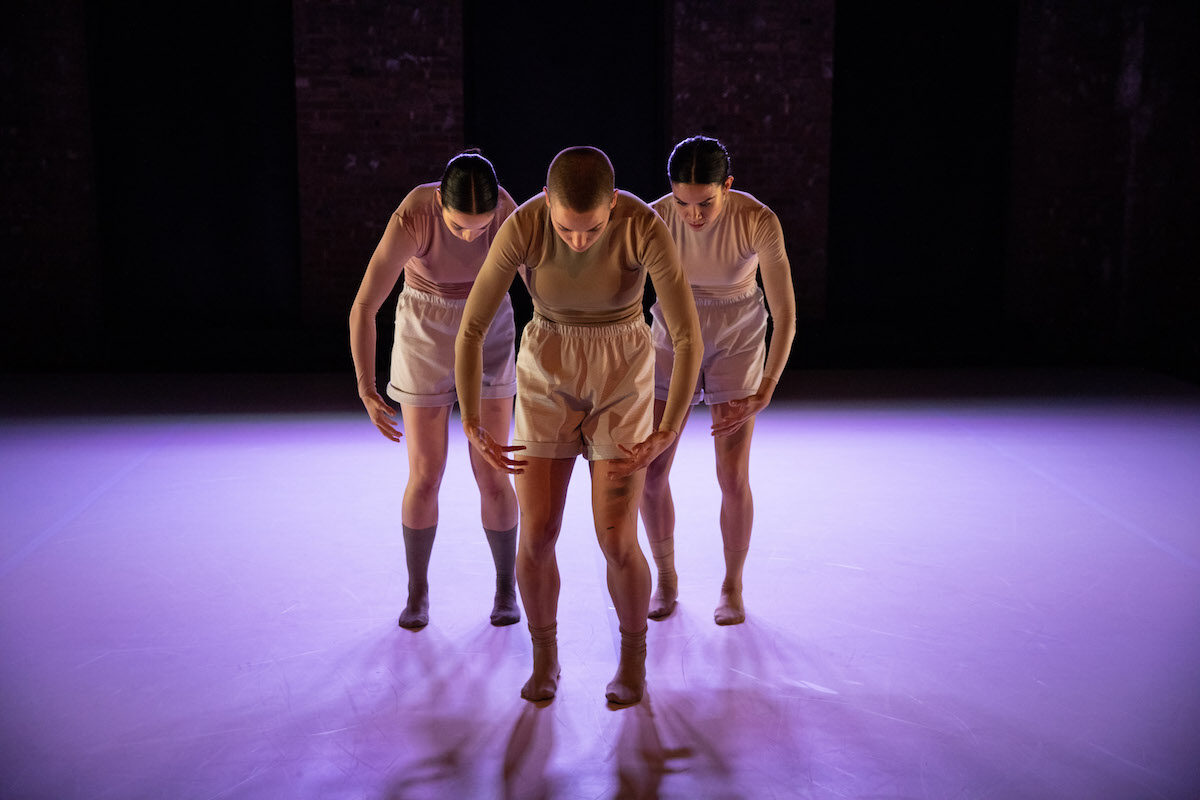 Three Gibney Company dancers standing in a V shape formation gazing down at a purple floor.