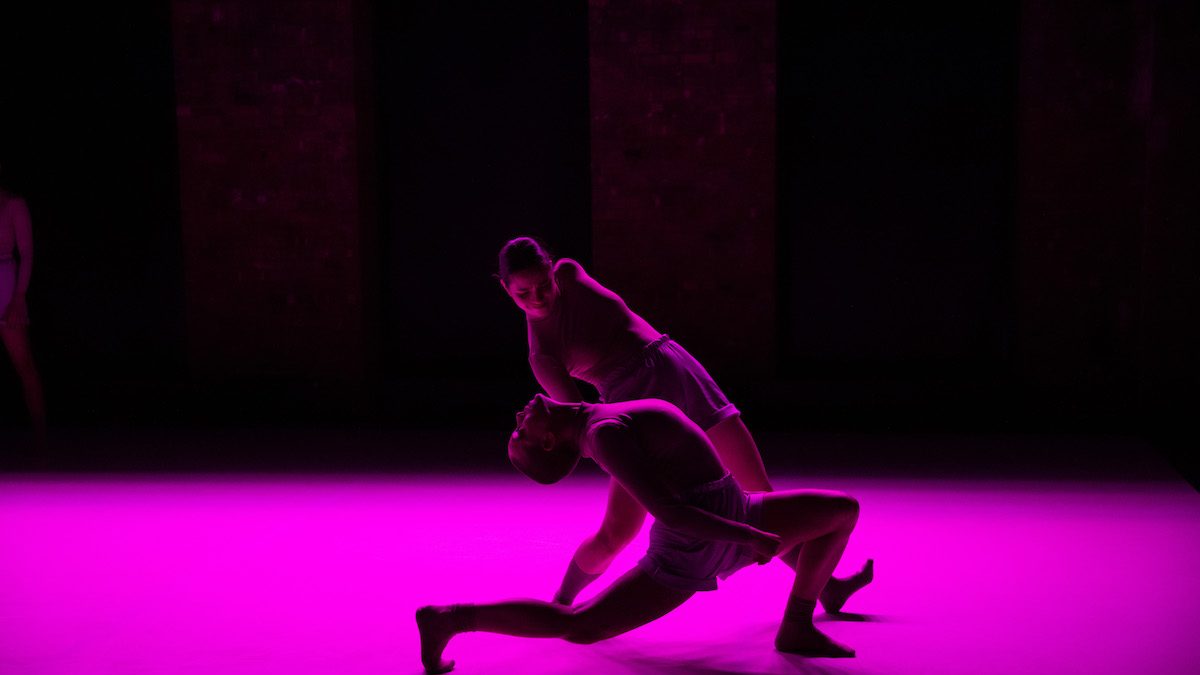 One woman holding another by her neck mid-motion on stage. Both are standing on a saturated pink floor.