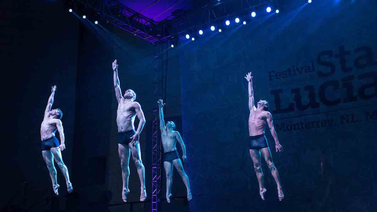 Group of men jumping in the air on stage with their right arms lifted high.