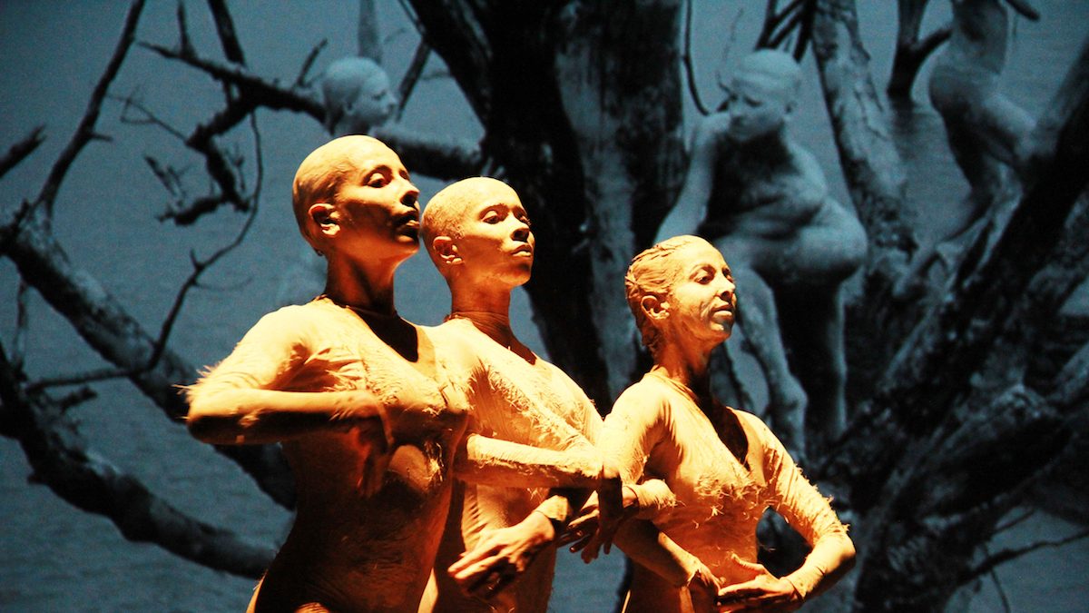 Three women on stage with painted bodies, linking arms.