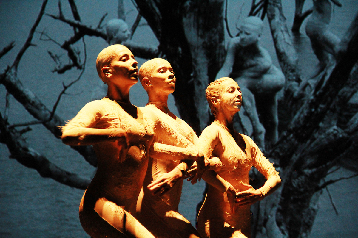 Three women on stage with painted bodies, linking arms.