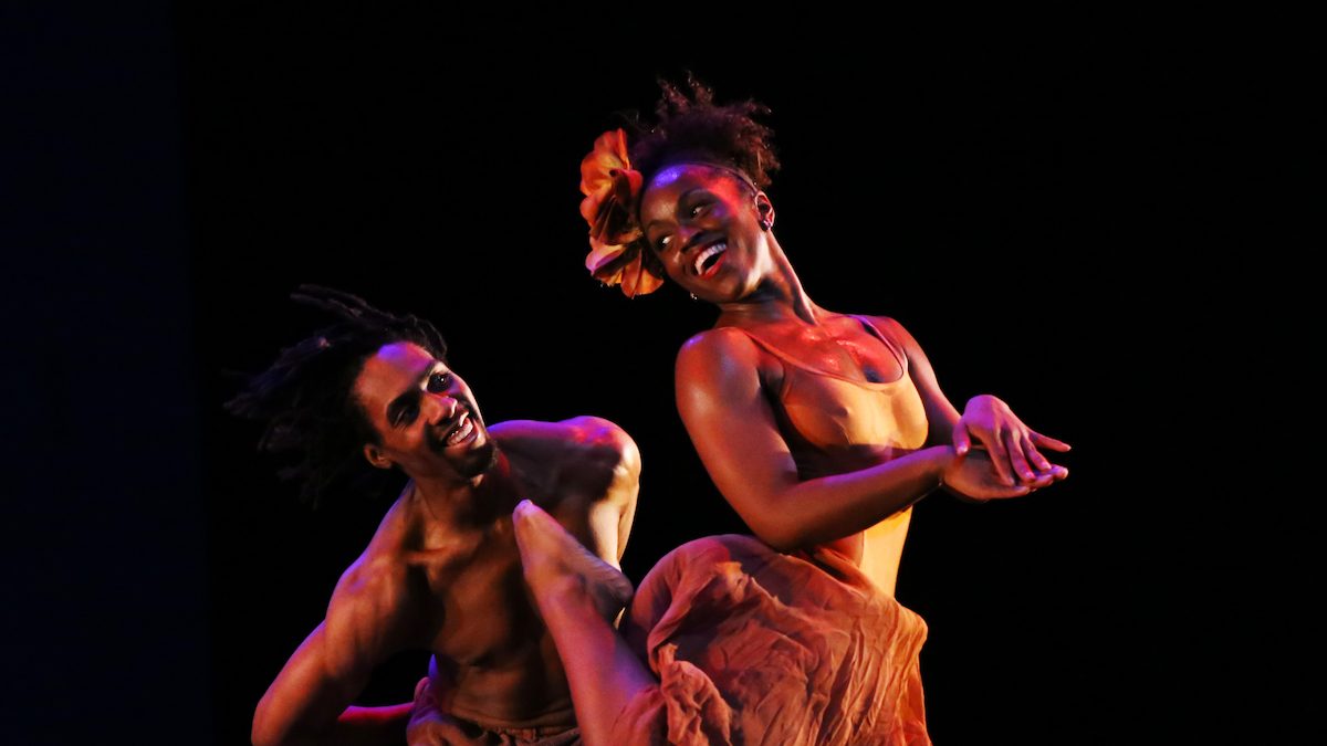 Two black dancers smiling and looking at each other on stage.