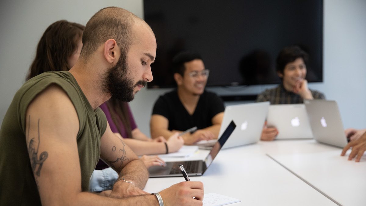 Man writing amongst a group speaking at a table in an office.