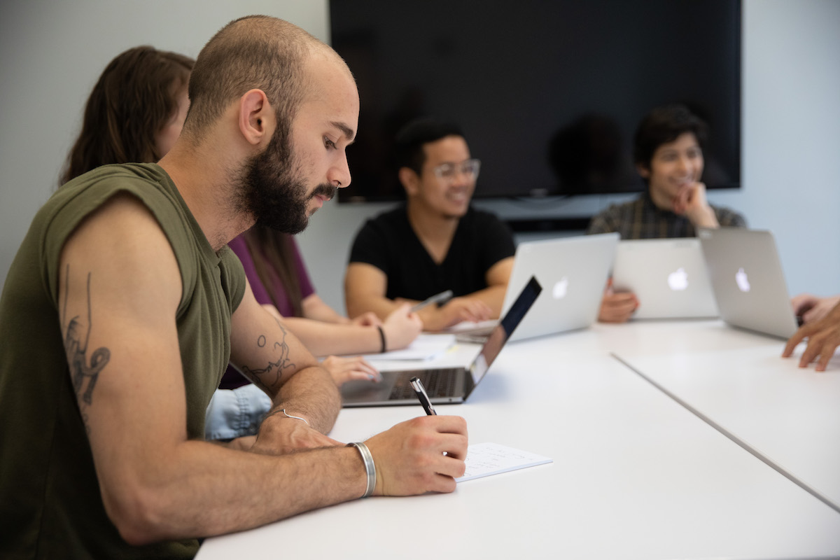 Man writing amongst a group speaking at a table in an office.