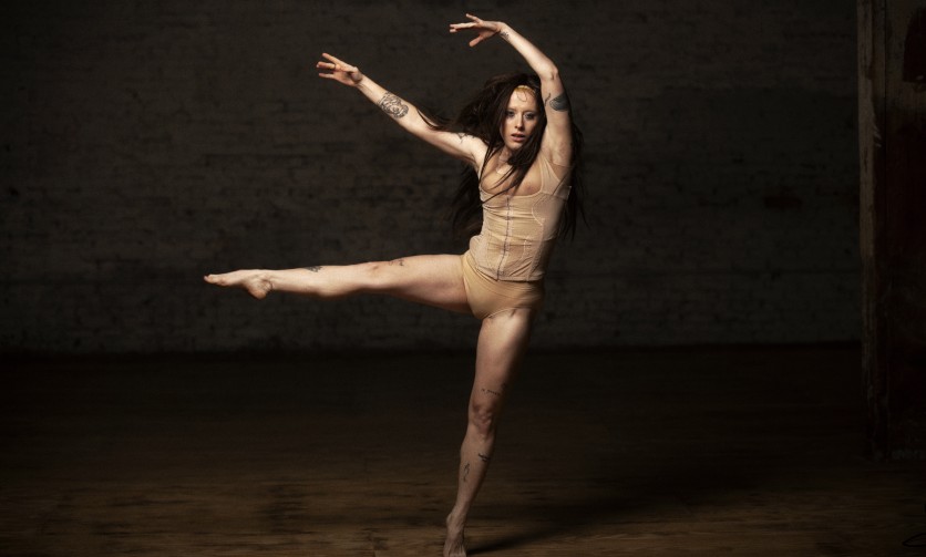 White woman in a ballet position, mid-motion in a nude suit.