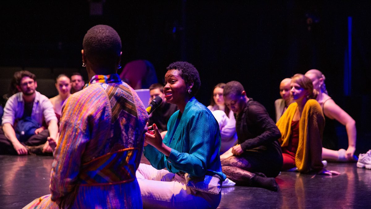 Black woman speaking to a group sitting in a circle with a microphone.