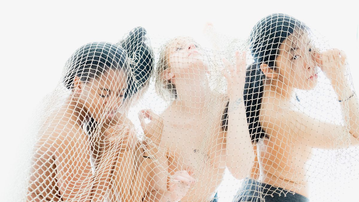 Four femmes topless, standing inside a net cloth.