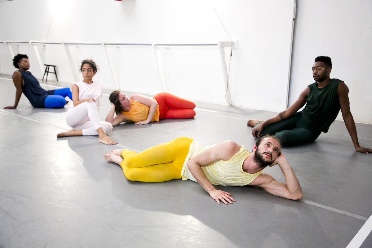 Group of dancers sitting in a configuration, each wearing solid colors.