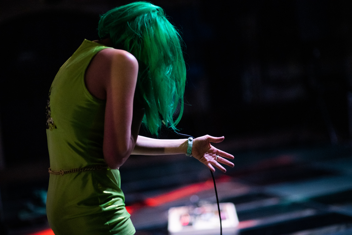Person wearing green with green hair, as they speak into a microphone.