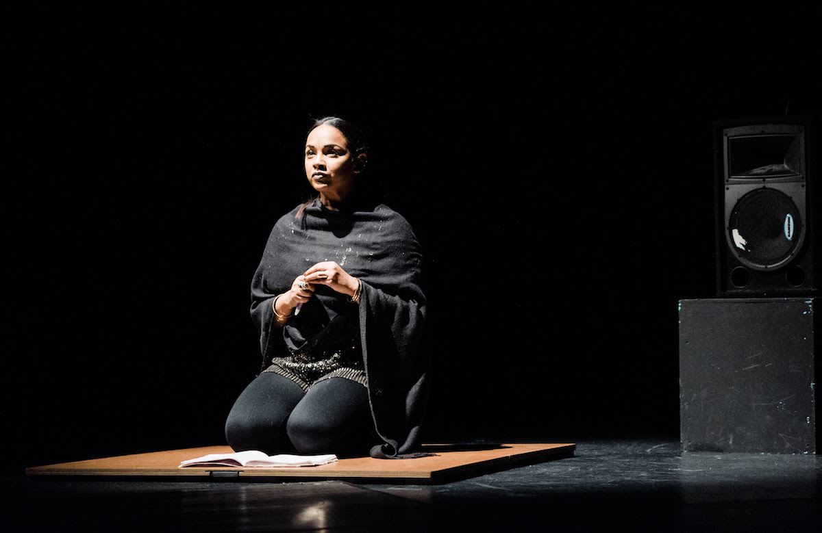 Woman of color dressed in black on stage, kneeling on the floor.