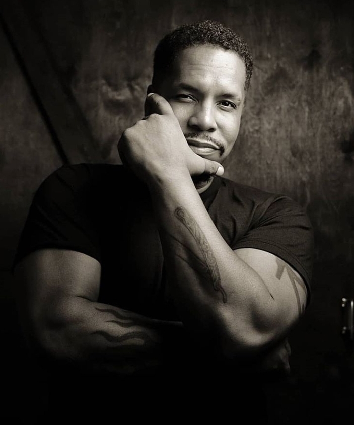 Black and white photo of a Black man smiling at the camera with his hand on his chin.