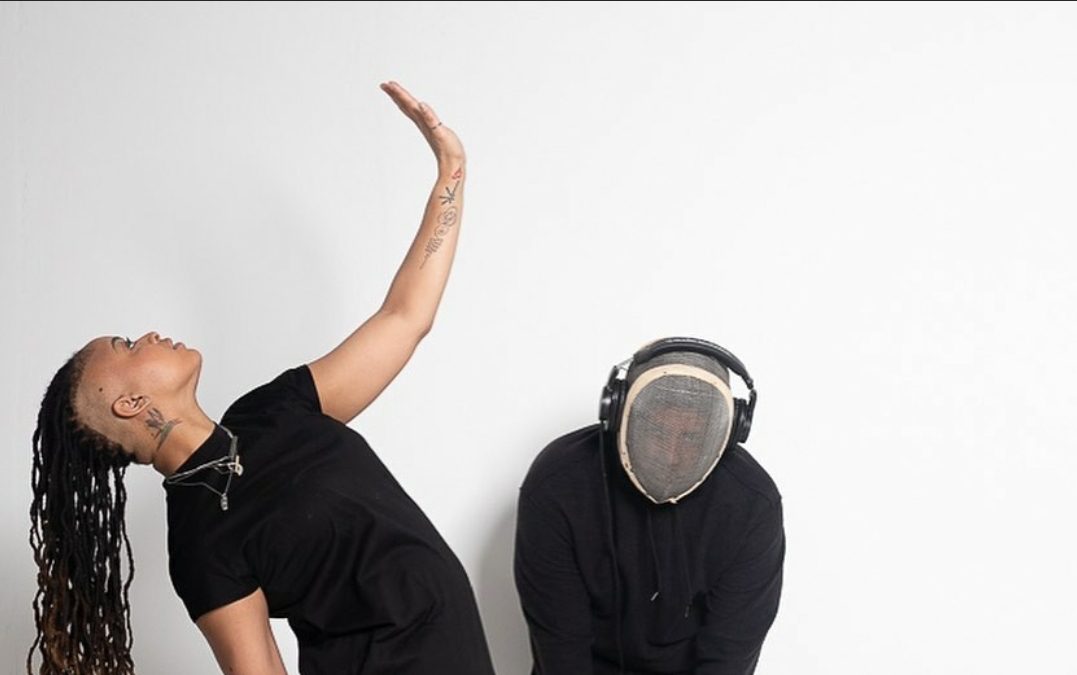 A Black woman and man behind her posing in a white studio.