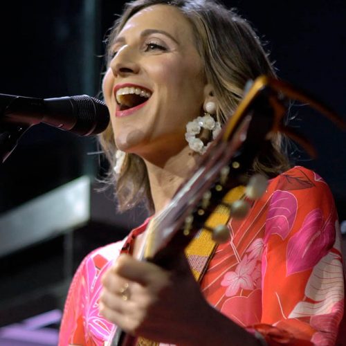 White woman playing guitar into a microphone.