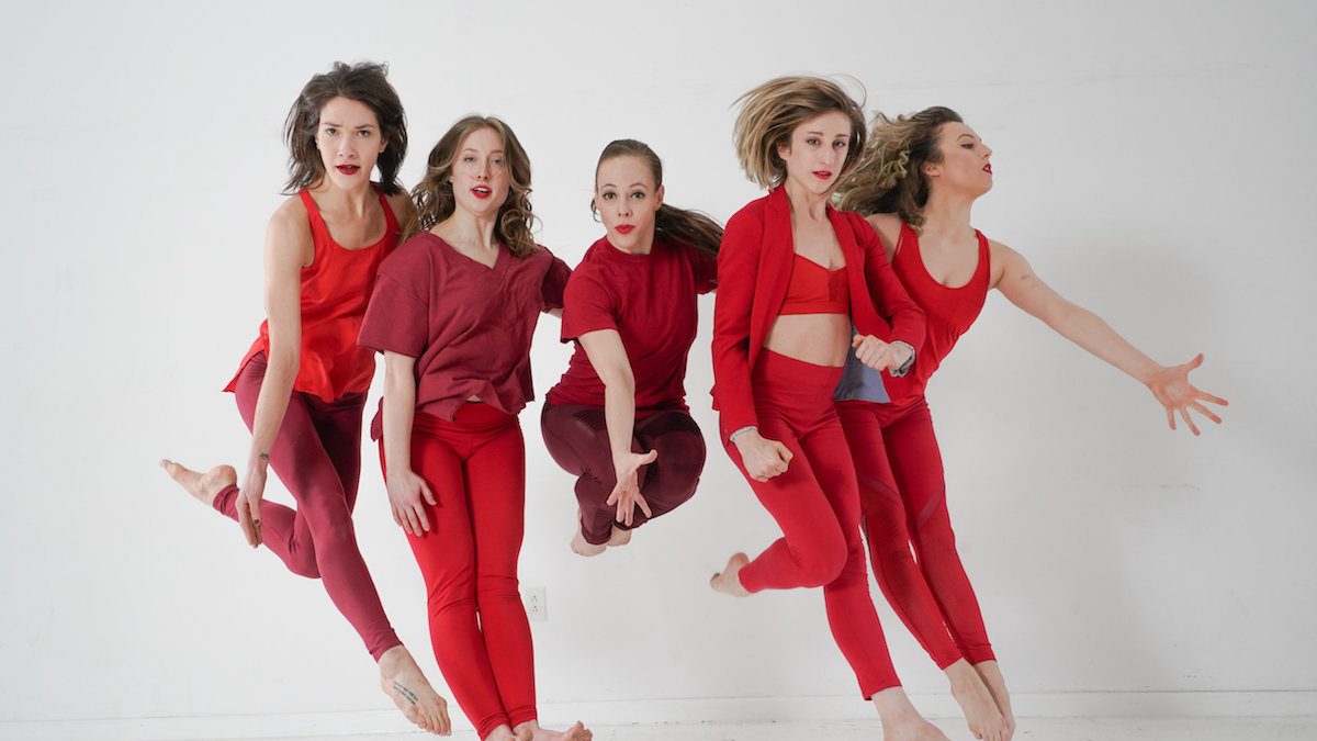 Five white women jumping in the air, wearing red.