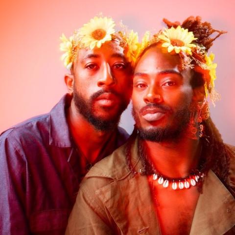 Two black men with sunflowers on their head looking at the camera.