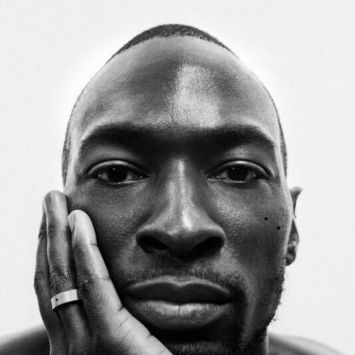 Photo of a Black man looking at the camera with his right hand resting on his face.