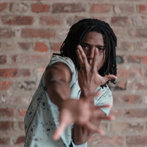 Black man dancing as his hands cover his face towards the camera.