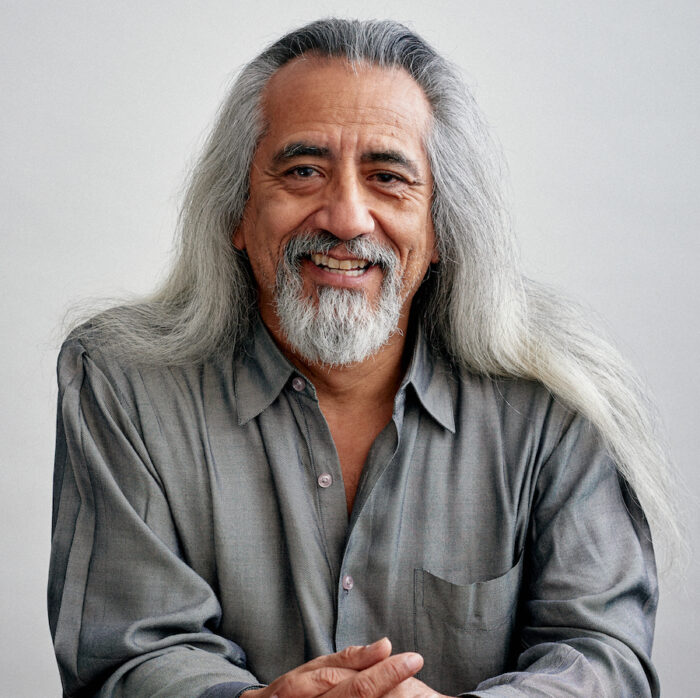 A man of color smiling at the camera with long grey hair and a grey shirt.