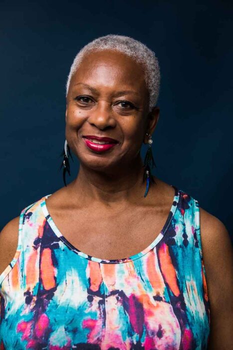 Photo of Eva Yaa Asantewaa by Scott Shaw • Image description: Eva Yaa Asantewaa (she/her) faces the camera against a teal-colored background. She is a Black woman with medium-brown skin, dark-brown eyes, and very short grey hair. She's wearing a multicolored, sleeveless dress with a scoop neckline and dangling earrings made of plastic feather-like pendants and pearls. Wearing cranberry-red lipstick, she's smiling warmly, her head tilted slightly towards her left shoulder.