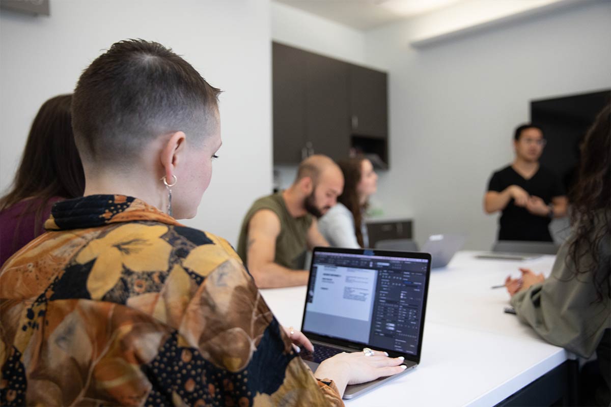 A woman with short dark hair sits at a computer working, with others in conversation around her. - Climate of Possibility