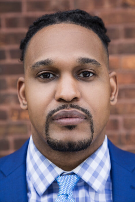 Headshot of Durell Cooper, wearing a blue and white plaid shirt, with at light blue tie, and a navy blue jacket, facing forward, behind a brick wall