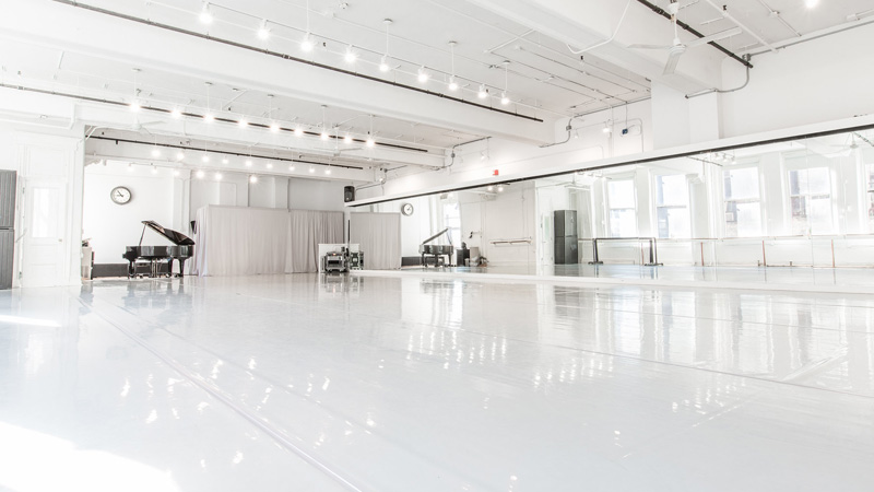 Photo of Studio 5-2 at Gibney 890 Broadway. A large, bright dance studio with white walls, a gray marley floor, large mirrors and a piano.