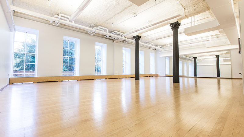 Photo of Studio D at 280 Broadway. A sunlit dance studio with wooden floors, white walls, a large wall of windows looking out on trees, and two gray columns.