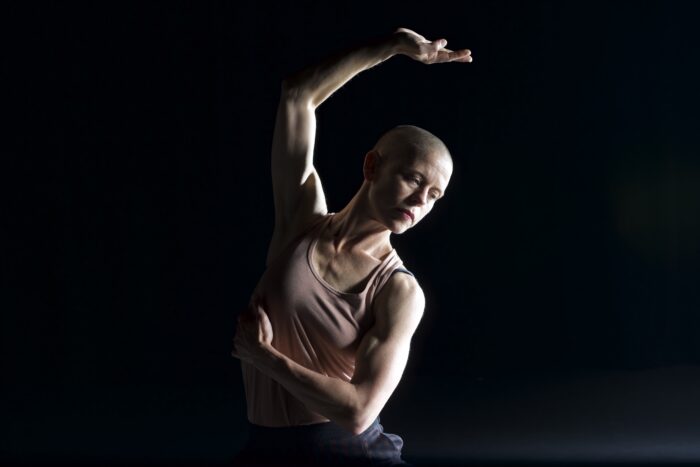 Jim Coleman movement photo, with one arm bent slightly towards the air above her head, and the other bent down across her chest, wearing a light beige, tank top.