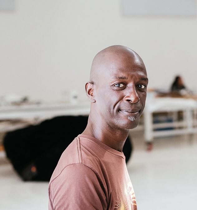A bald clean shaven dark-skinned man, David Thomson, with a salt and pepper soul patch, in a large white studio, wearing a brown tee shirt.
