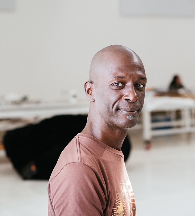 A bald clean shaven dark-skinned man, David Thomson, with a salt and pepper soul patch, in a large white studio, wearing a brown tee shirt.