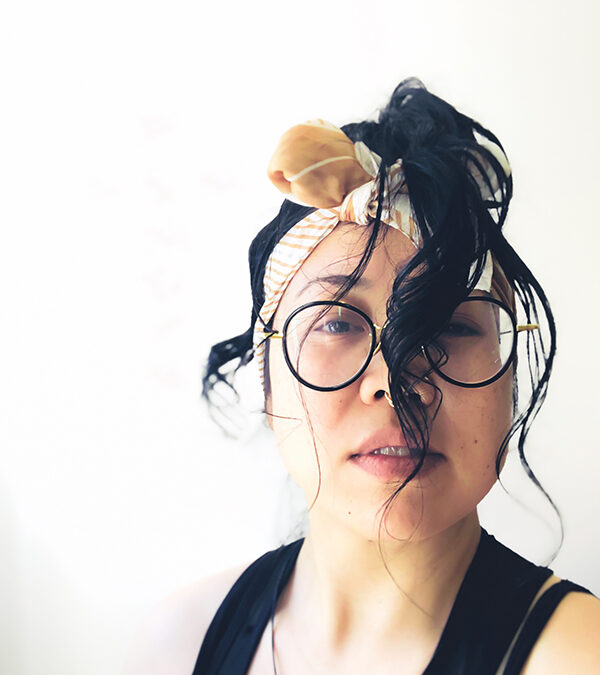 a headshot of día, a queer Vietnamese with jungle light, yellow power pigmented skin with mid-length black hair tied up in a pony-tail dangling in front of face and a white/tan headscarf tied like a head-band. día is wearing glasses with oddly round black rims and gold trimmings. día is wearing a gold septum and crystal nose rings with a black tank top and a simple black necklace with an ancient symbol of virility (lingling-o) from the Philippines that's out of frame.