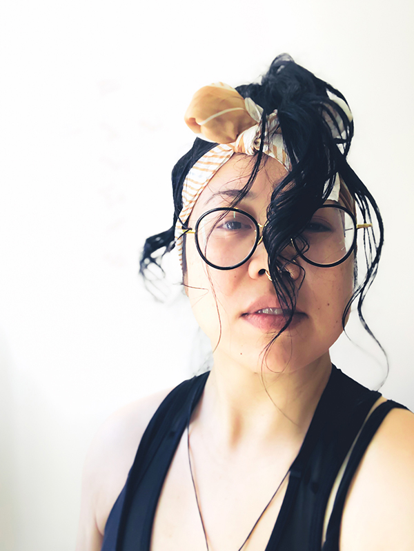 a headshot of día, a queer Vietnamese with jungle light, yellow power pigmented skin with mid-length black hair tied up in a pony-tail dangling in front of face and a white/tan headscarf tied like a head-band. día is wearing glasses with oddly round black rims and gold trimmings. día is wearing a gold septum and crystal nose rings with a black tank top and a simple black necklace with an ancient symbol of virility (lingling-o) from the Philippines that's out of frame.