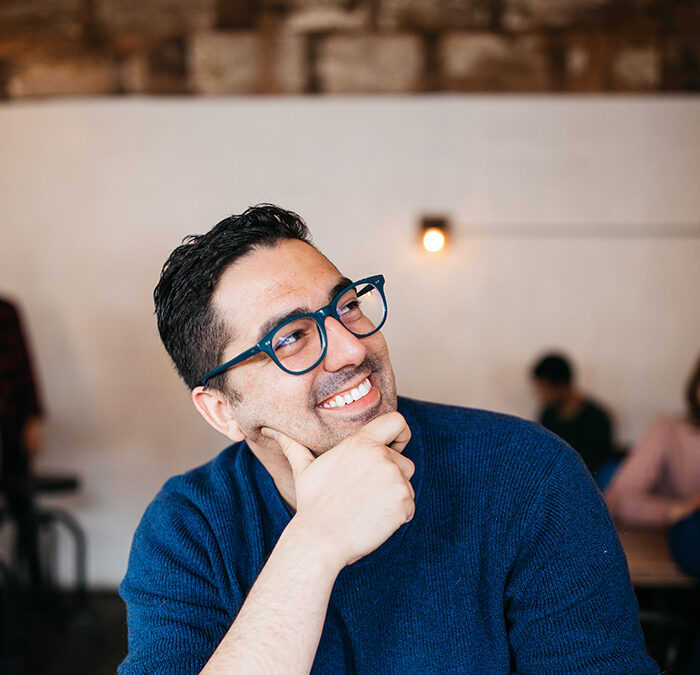 Greg is a white latinx man with short dark hair and glasses. He is looking up to the right with his hand on his chin. He is sitting in a coffee shop with half drank cup of coffee in front of him. He is wearing a blue sweater and has big smile on his face.