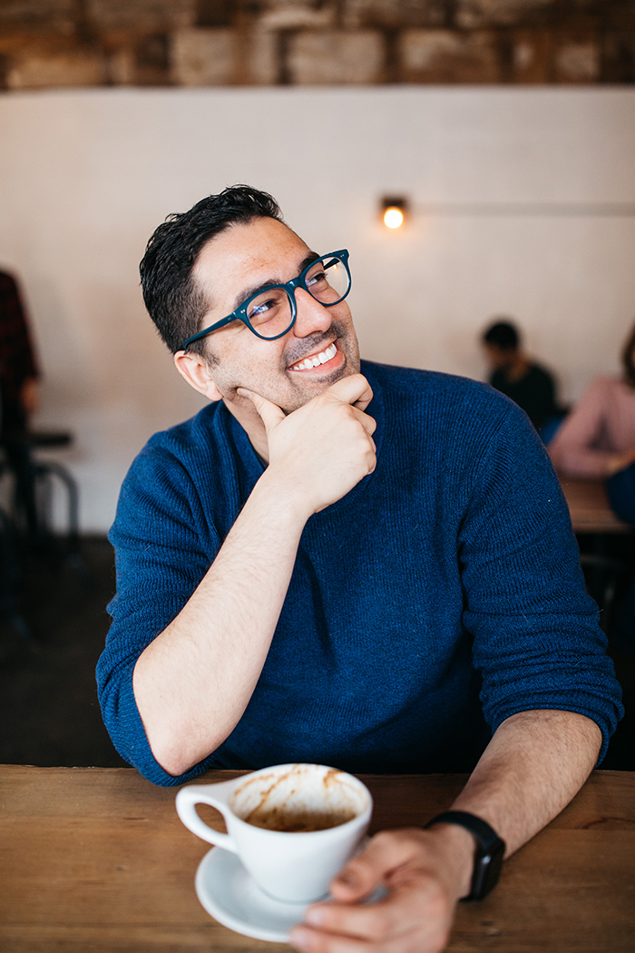 Greg is a white latinx man with short dark hair and glasses. He is looking up to the right with his hand on his chin. He is sitting in a coffee shop with half drank cup of coffee in front of him. He is wearing a blue sweater and has big smile on his face.