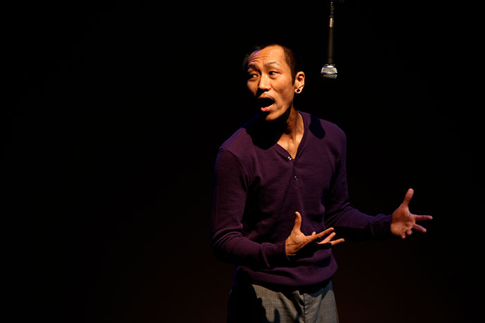 Makoto Hirano, a person of Asian descent in purple shirt; arms bent at elbows with hands open and palms facing slightly toward each other/up; a mic hangs upside down and Makoto’s mouth is open as if speaking or making sounds