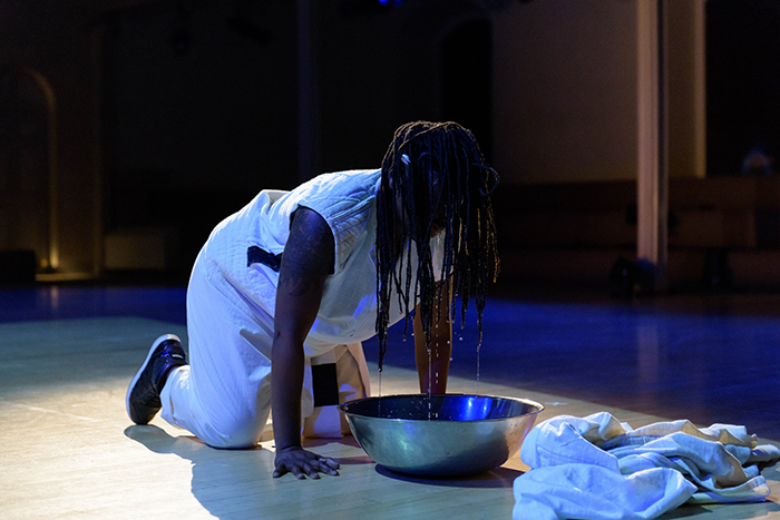 A deep-skinned trans artist wearing white is on their hands and knees in front of an aluminum bowl. Their hair drapes over their face and drips water into the bowl.