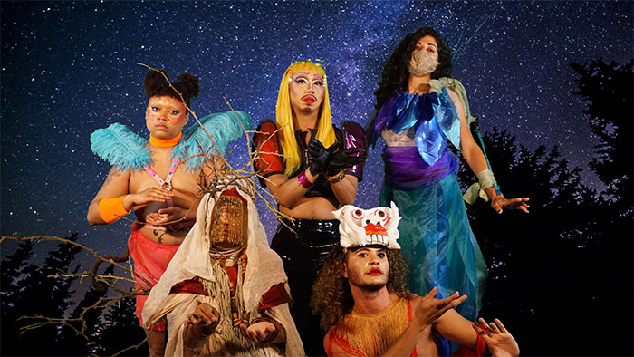 Five deities in colorful costume, masks, and drag look into the camera lens and pose boldly. Behind them is a digital backdrop of a starry night sky.