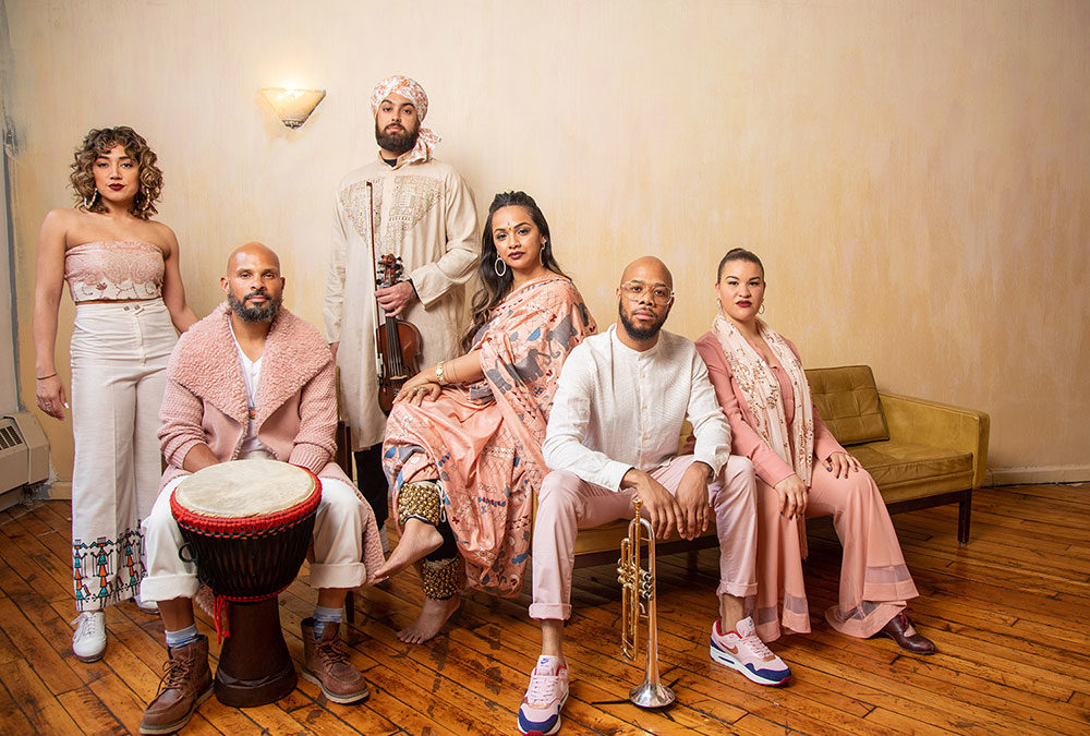 From left to right: Amanda (standing), Okai (sitting), Raaginder (standing), Brinda (sitting), Ryan (sitting), and Arielle (sitting) are gathered in diverse cream and rose gold outfits on a wooden floor and a mustard couch against a cream wall. Amanda has on a light pink tube top with a wide legged white with pattern pants and white tap shoes. Okai has on a light pink furry sweater with white pants, brown shoes, and a wide-rimmed djembe sitting in front of him. Raaginder has on a cream tunic and a white and pink floral turban and is holding a violin and bow. Brinda is draped in a light pink (with stitch pattern) sari and is wearing ghungroo ankle bells and bare feet. Ryan is wearing a full-sleeved white top and light pink pants with pink sneakers and a trumpet propped on a stand in front of him. Arielle is wearing a full pink outfit and light pink scarf around her neck as well as maroon flamenco shoes. All performers are looking at the camera with calm and focused facial expressions.