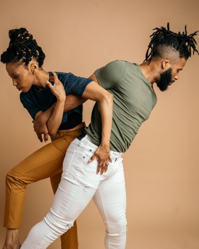 Photo by BeccaVision alt text image description: tan background, artistic directors Latra Wilson (L) and Winston Dynamite Brown (R) locked in a mid-stride partnered stance glancing away from each other.
