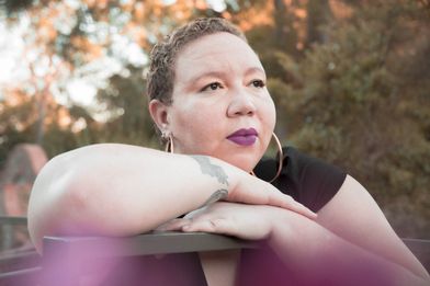 Photo by Janae Brown light skinned black woman with sandy colored hair with blondish highlights, purple lipstick and gold hoop earrings. She has on a black top, tattoos around her right wrist, and her arms are folded leaning onto a black bar with her chin rested on her hands looking out.