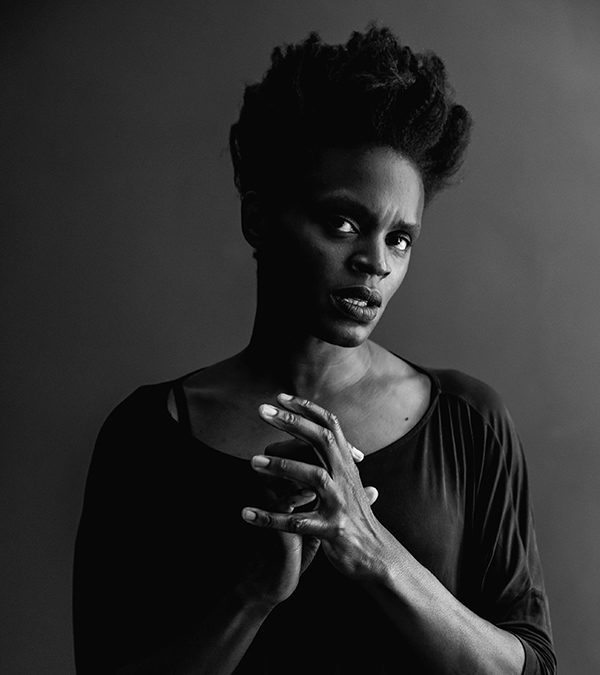 In this black and white image, Okwui is wearing a loosely fitting dress. Her elbows are along the sides of the body and her forearms reach towards each other in the center, connected but not clasped. Okwui is not smiling but looking up and with a side glance. Her hair is black and in a shaped Afro standing high on top of her head.