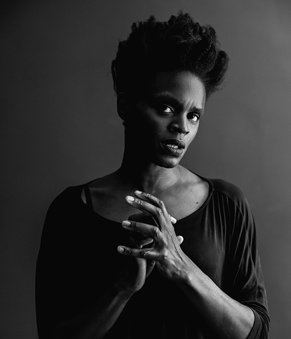 In this black and white image, Okwui is wearing a loosely fitting dress. Her elbows are along the sides of the body and her forearms reach towards each other in the center, connected but not clasped. Okwui is not smiling but looking up and with a side glance. Her hair is black and in a shaped Afro standing high on top of her head.