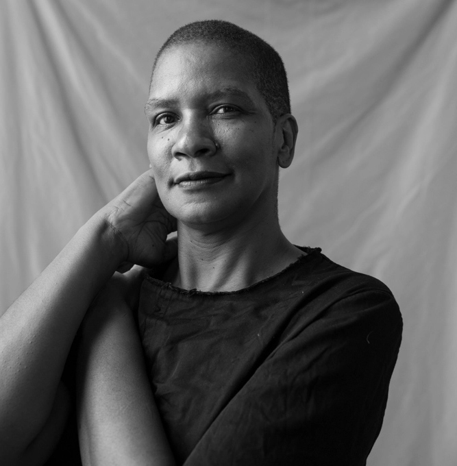 Pamela Sneed is a Black woman with close-cropped hair. In this cropped black and white photo, she stands in front of an elegant gray cloth backdrop, looking directly at the camera. Pamela is wearing a black shirt with one arm folded across her chest. She holds the side of her neck with the other hand.