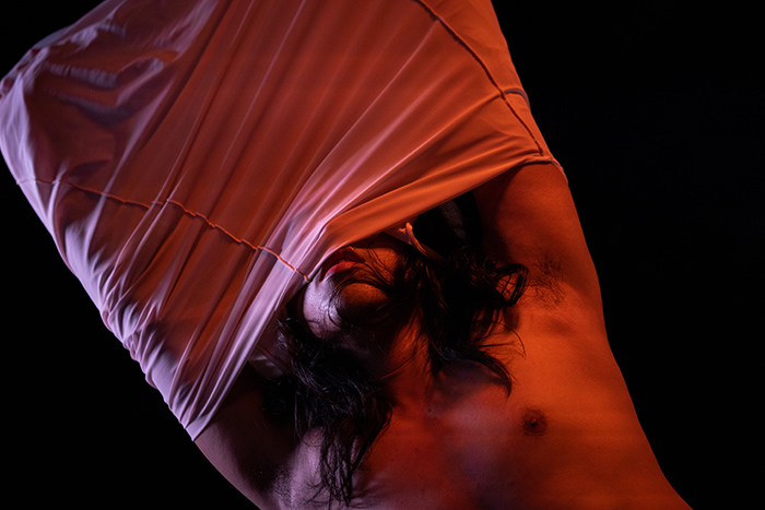 This photo shows performer Anh Vo in the middle of pulling a shirt off, and so the shirt is inside out, its seams showing. The performer's arms are lifted above their head, encased in the fabric of the shirt which partly stretches across and covers most of their face. Vo's lips and chin remain uncovered. Dark hair spills down from around their head. Vo's lower torso is also uncovered, the left nipple shown. This image is drenched in red light and is set against a dark background.