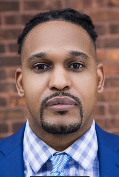 handsome Black man wearing a blue and white checkered pattern button up shirt with the top button open and a solid blue jacket over it against a brick background.