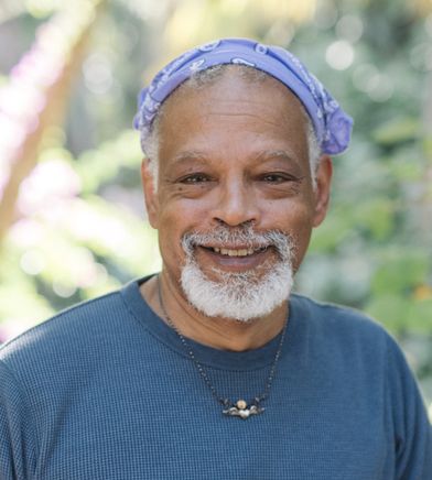 Ishmael, and African American man with a lavender bandana and a gray goatee, is smiling outside on a bright day, wearing a blue shirt and a winged-heart necklace. Behind him is a blur of trees.